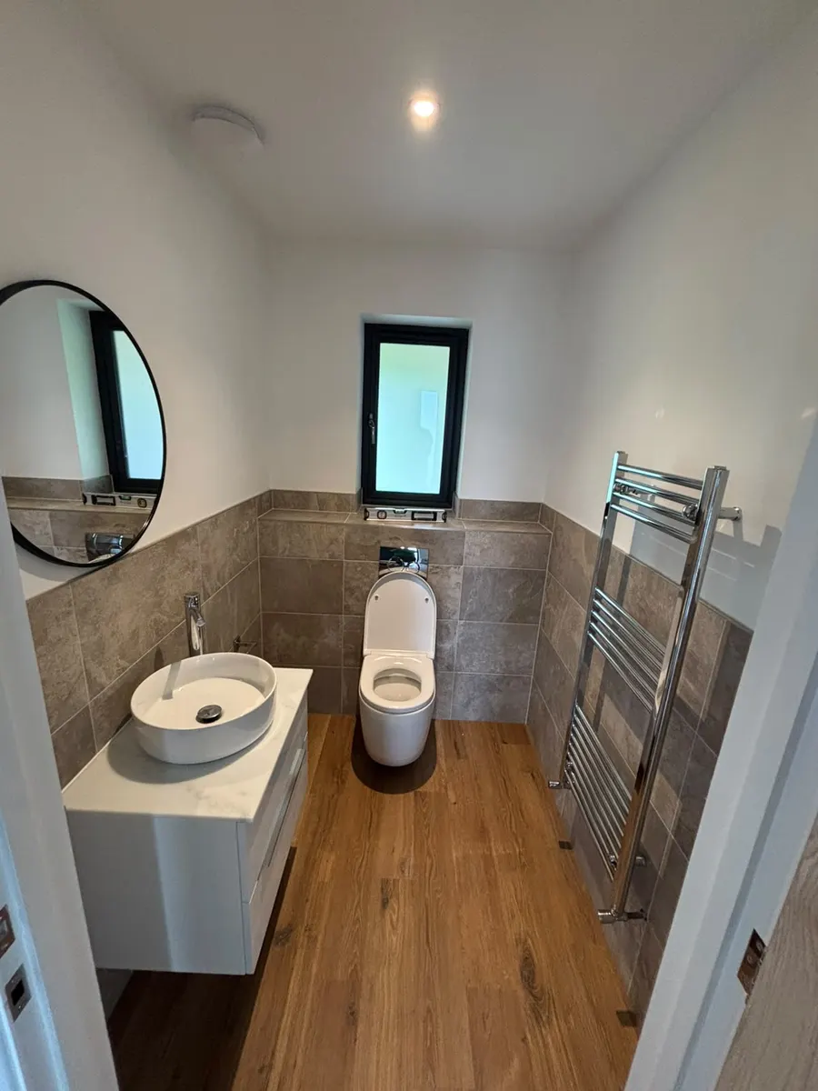 Finished bathroom with wall-hung toilet, round countertop basin on a white floating vanity, round black mirror, half-height grey stone-effect tiles, chrome towel rail and oak-look floor